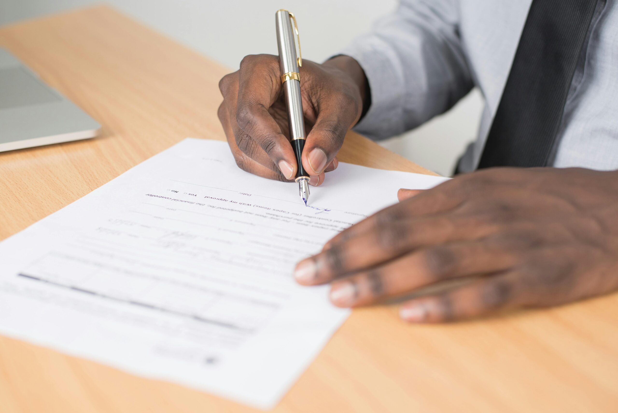 Person signing an official document with a silver pen at a wooden desk.