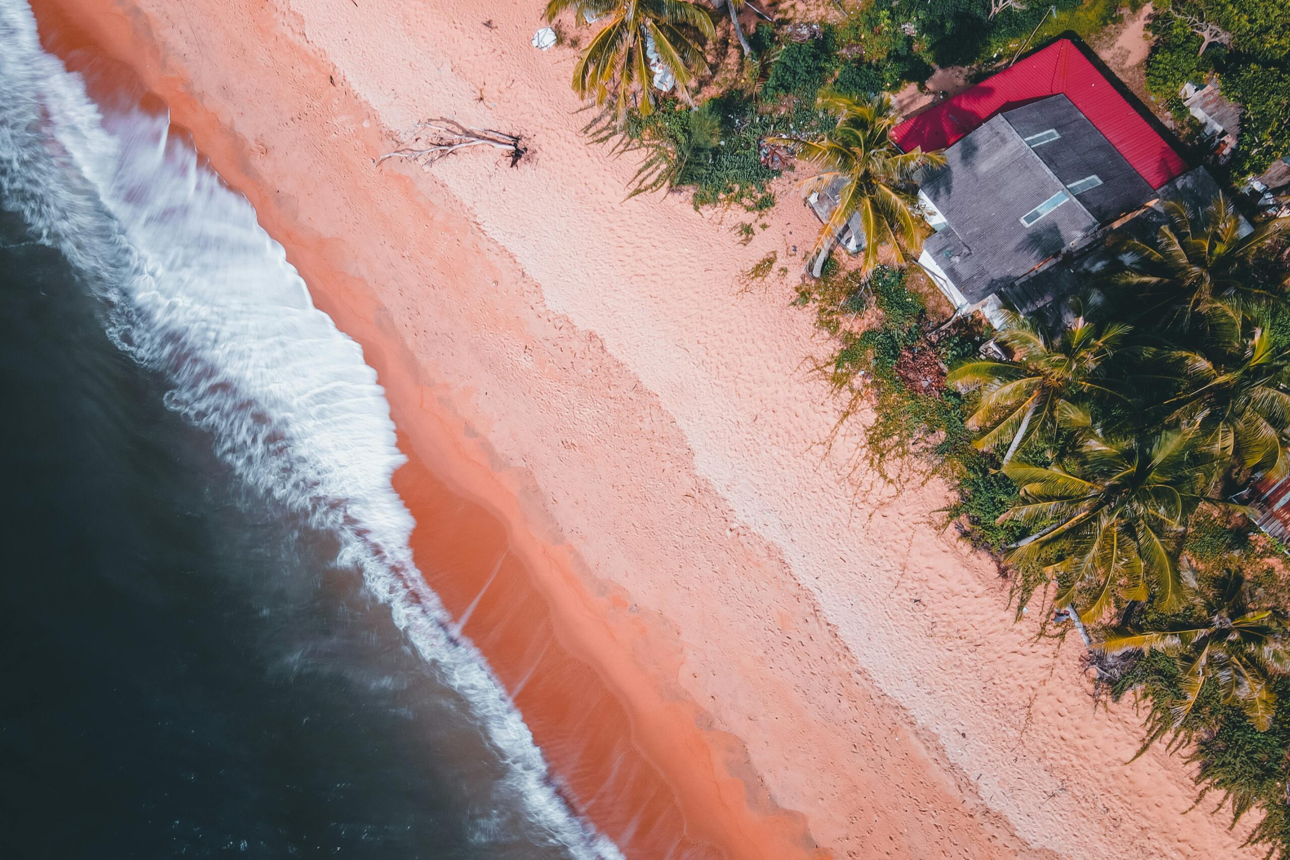 Aerial view of waves hitting a tropical beach with palm trees and a red-roof house nearby.